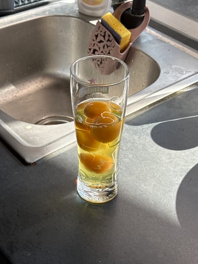 A close-up shot captures a glass of amber liquid with whole plums submerged within it, resting on a dark countertop. The glass, adorned with faint lettering and a small star, sits to the right of a stainless steel sink. In the background, a pink dish rack holds a yellow sponge and a purple scrubber, positioned near a faucet. Sunlight casts sharp shadows across the scene, highlighting the textures of the countertop and the sink. The image appears to be taken from a slightly elevated perspective.