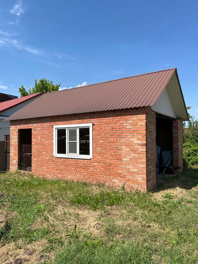 This is a full-shot, eye-level, outdoor, daytime, full-color photograph of a modest brick garage in the city of Takhtamukai, Russia. The garage, constructed of orange-red brick, has a brown corrugated metal roof and a white gable end. A white-framed window with three panes is visible on the front facade. The left side of the garage has an open doorway with a metal grate. The right side also has an open doorway, revealing stored items inside, including what appears to be a green tank and pipes. The structure is surrounded by dry and green grass. In the background, trees and the corner of another building with a red roof can be seen under a clear blue sky with scattered white clouds. There are no people in the image.