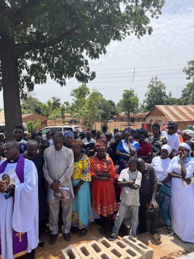 The image depicts a gathering of people in Dogon Dinia, Nigeria. A large group of people of varying ages are assembled outdoors, many of whom are seated. The people are dressed in a mix of traditional African clothing, casual attire, and religious garments. A man wearing a white and purple clerical collar appears to be a religious leader, holding what looks like printed materials. The gathering is taking place near buildings with corrugated metal roofs, and a parked white van is visible in the background. A large tree stands on the left side of the frame. The weather appears to be clear and sunny.