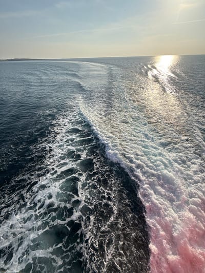 The image displays an expansive view of the Aegean Sea near Kamariotissa, Greece, observed from the stern of a moving vessel. The central subject is the vessel's wake, a prominent, frothy trail of white and light blue churned water originating from the bottom right and curving diagonally towards the center-left horizon. To the left of this turbulent wake, the sea water is a darker blue-green, exhibiting patterns of less disturbed surface foam. A low-angle sun, positioned in the upper right quadrant, casts a brilliant, elongated reflection path across the water, particularly illuminating the right side of the wake with a silvery-gold shimmer. A subtle pinkish tint is present within the brightest foam at the bottom right. The horizon line is distinct and level, separating the deep blue sea from a pale blue sky with minimal cloud cover and a faint contrail in the upper right. A low, dark landmass is discernible on the far left horizon, indicating proximity to a coastline or island. No individuals or specific objects are identifiable apart from the natural elements and the implied vessel creating the wake.