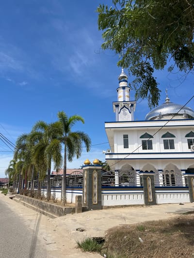 A mosque with a dome and minaret is pictured on a sunny day in Dalam, Indonesia. The mosque is white with blue and gold accents. Palm trees line the street in front of the mosque, and power lines are visible overhead. The sky is blue with some scattered clouds. The ground in front of the mosque is a paved walkway and a dirt area with dry grass. There is a stone wall in front of the mosque with decorative pillars.