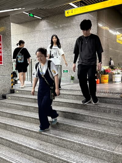 Here is a forensic description of the image.

The image depicts an indoor scene, seemingly inside a subway station in the 龙溪街道 area of China. A group of four individuals is present on a set of stairs. One person wears all black clothing, another wears a white shirt and jeans, and a third person is wearing a white shirt with dark pants. The final person is wearing a black shirt and shorts. A yellow sign with Chinese characters and the number 4 is visible, likely indicating an exit ("出口") towards Honghuang Road (红黄路). Various signs are displayed in the background, including emergency exit signs and potentially advertisements. The presence of stairs suggests the location is not on ground level, but the overall details indicate a relatively clean and modern public space.