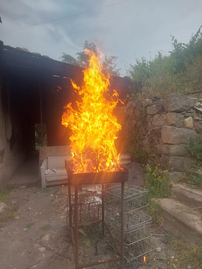 A bright, roaring fire burns in a metal grill set on a stand in an outdoor, rural-looking area. The fire is the central and most dominant element of the image, with flames reaching high into the sky. To the left of the grill, a covered structure with a dark, open doorway is visible. Inside the structure, a light-colored sofa with some white fabric on it can be seen. To the right of the grill, a rough stone wall is present, overgrown with greenery and small yellow flowers. The ground is dirt and gravel, with scattered rocks and dry vegetation. Two stacked wire baskets are positioned to the right of the grill stand. The overall scene suggests a rustic or semi-abandoned outdoor setting, possibly a backyard or a less-maintained area. The sky is overcast with a light gray hue. This location is consistent with the appearance of a residential property in the village of Areni, Armenia, given the rustic architecture, stone walls, and vegetation.