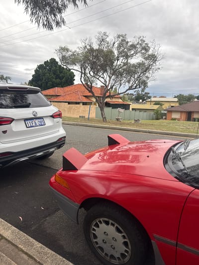 The image depicts a street scene in the city of Bassendean, Australia. A white MG SUV is parked behind a red car with pop-up headlights. The license plate on the MG reads "1IFA285". The vehicles are on a paved street next to a concrete curb. In the background are houses with tiled roofs, a tree with bare branches, and a cloudy sky.