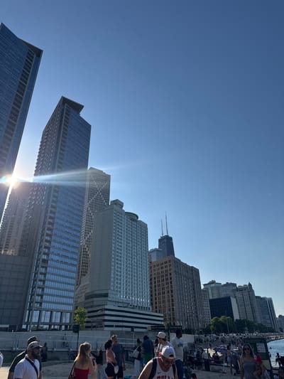 The image shows a view of the skyline of Chicago, Illinois, with several tall buildings visible against a clear blue sky. The sun is shining brightly, creating lens flare effects. People are situated near the waterfront, with some sitting and others walking. The John Hancock Center is visible among the buildings. The scene appears to be from near Ohio Street Beach on a sunny day.
