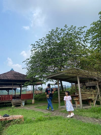 A young girl stands near a man outside of a rustic, partially enclosed wooden structure with a tile roof. The girl wears a light-colored dress and white shoes. The man wears a dark t-shirt, blue jeans, and casual shoes. They appear to be located in a grassy area near a path. The setting appears to be Penanggungan, Indonesia. A large tree provides shade in the foreground, and additional similar structures are visible in the background. The sky is visible above.