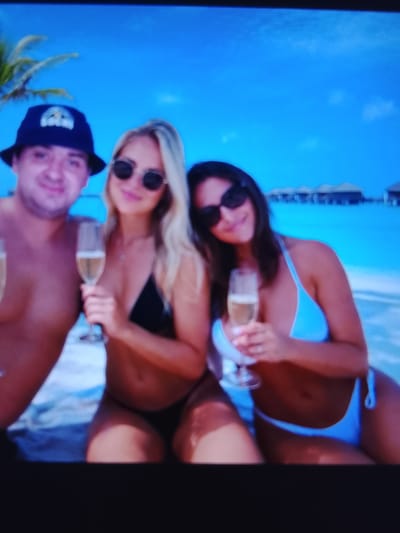 The image shows three people on a beach with a tropical ocean in the background. On the left, a man wearing a dark bucket hat and no shirt smiles at the camera. Next to him, a woman in a black bikini holds a glass of champagne and wears sunglasses. To her right, another woman in a light blue bikini also holds a glass of champagne and wears sunglasses. In the background, there are overwater bungalows, a palm tree, and the ocean. The location is not Safonovo, Russia.