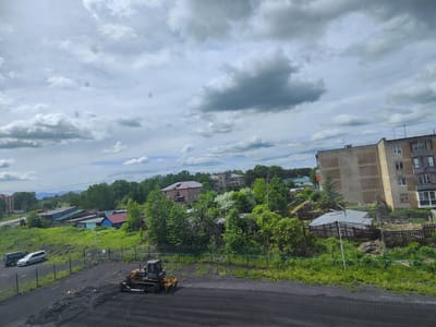 The image captures a scene in Milkovo, Russia, on a cloudy day. In the foreground, a bulldozer is parked on a dark, dusty surface, likely coal or ash, with tire tracks visible. A green fence runs horizontally across the middle ground. Beyond the fence, several vehicles are parked, including two SUVs, one dark and one silver. Further back, a collection of low-rise buildings with various colored roofs (red, blue, green) are nestled amongst lush green trees. In the background, a tall, multi-story building, typical of Soviet-era apartment blocks, dominates the right side of the frame. The sky is filled with dramatic, grey clouds, hinting at potential rain. The overall impression is one of a rural or semi-urban environment undergoing some form of construction or maintenance.
