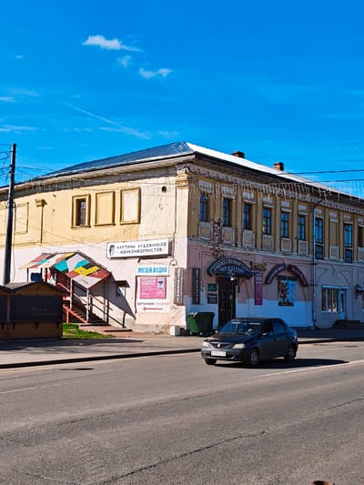 The image shows a two-story building in Uglich, Russia. The building has a mix of yellow and pink exterior paint and a metal roof. Signs on the building advertise an art gallery, a vodka museum, a chocolate shop, and Uglich watches. A dark gray car is driving on the road in front of the building. There is a green dumpster next to the building. A small, covered structure with colorful panels on its roof sits to the left of the main entrance. The sky is blue with a few scattered clouds.