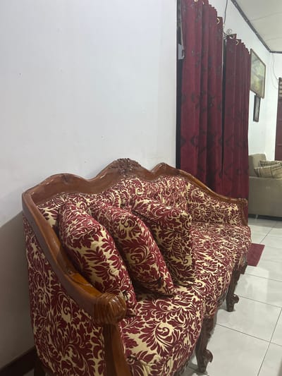 The image shows an ornate sofa with a dark red and cream floral pattern, set in a brightly lit indoor space. The sofa has a carved wooden frame with a warm, polished finish. Multiple plush cushions are arranged on the sofa, creating a comfortable and inviting appearance. In the background, several burgundy curtains with a geometric pattern are visible, hung from a rod with gold grommets. A framed picture hangs on the wall above the curtains. To the right of the sofa, the wall is tiled with white square tiles. The scene appears to be a room in a home or a furniture showroom, likely during the daytime given the lighting. There are no people visible in the image, and the focus is entirely on the furniture and its surroundings. The overall impression is one of elegance and comfort.