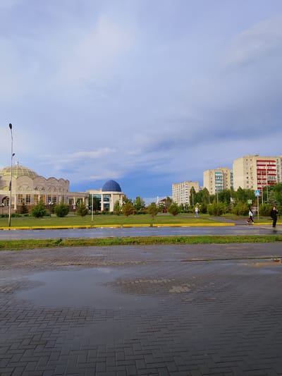 This image depicts an urban scene in Oral, Kazakhstan, following a rain shower. The sky is overcast with dramatic clouds, suggesting either early morning or late afternoon. The wet pavement in the foreground reflects the sky and surrounding environment, indicating recent rain.
The scene features several buildings of varying architectural styles. To the left, a large, curved building with a glass dome and ornate detailing is visible, possibly a cultural center or a government building. Adjacent to it, on the right, is another building topped with a bright blue dome, standing out against the muted sky. In the background, a row of tall, modern apartment buildings with balconies and red and white accents dominates the skyline. Green spaces with trees and manicured lawns are interspersed between the buildings, suggesting a planned urban development.
Several people are present in the image, walking across the street or on the sidewalks. Their presence suggests everyday activity, unaffected by the recent weather. A street lamp stands tall on the left side of the frame, a common urban fixture. There are no visible signs with text. The overall mood is calm and somewhat subdued, likely due to the weather conditions. The reflections in the puddles add a sense of depth and atmosphere to the image.