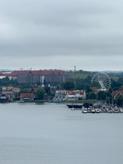 This media file captures a waterfront scene in Mikołajki, Poland. The image, oriented vertically, showcases a wide expanse of water on the right, with a cityscape lining the left bank. The cityscape is a mix of buildings, including a large, multi-story hotel with a dark red roof dominating the mid-ground. Adjacent to it, there are smaller buildings with red-tiled roofs, some featuring visible solar panels.
In the background, a Ferris wheel stands prominently, suggesting a recreational area or amusement park. A construction crane is also visible, hinting at ongoing development or maintenance. Along the waterfront in the foreground, numerous boats are docked, indicating a marina or harbor.
The sky appears overcast, suggesting a cloudy day. The water is calm, reflecting the muted tones of the sky. The overall impression is one of a coastal town or resort, likely during daylight hours given the visibility of the scene. There is some text visible on a building near the water's edge, appearing to be "Port de...". Another sign near the bottom of the image reads "BOAT RENTAL 874216163".