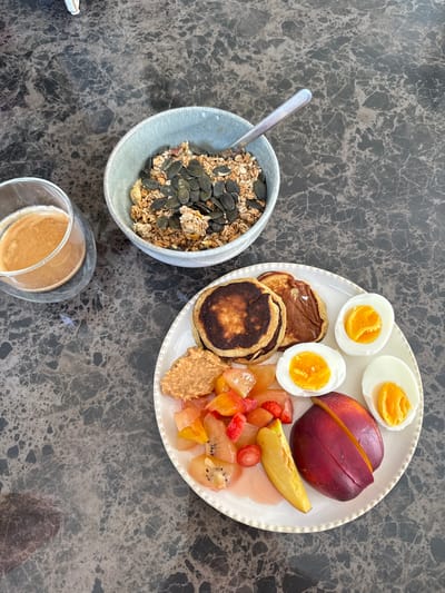 This is a top-down, eye-level medium shot of a breakfast spread on a dark marble countertop in Rueil-Malmaison, France. The breakfast includes a bowl of granola with pumpkin seeds, a glass of coffee, and a plate of food. The plate contains three small pancakes, a dollop of peanut butter, a fruit salad with kiwi, strawberries, and possibly peaches or nectarines, a halved nectarine, and three halves of hard-boiled eggs with bright yellow yolks. A spoon is resting in the granola bowl. The overall scene suggests a healthy and appealing morning meal.