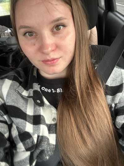 A young woman with light brown hair is seated in the driver's seat of a car, wearing a black and white checkered shirt over a black t-shirt. She is looking at the camera, with a seatbelt visible across her chest. The background shows the interior of the car and a blurred outdoor scene. There are no discernible landmarks or elements within the image that would allow for identification of the specific location within Kvarsa, Russia.