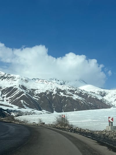 The image depicts a winding asphalt road through a snow-covered mountain landscape in Kanobi, Georgia. The road, appearing dark and clear of snow, curves sharply to the right in the foreground, with its left edge extending out of frame and its right edge bordered by a low, rough stone wall and sparse, leafless bushes. Two white rectangular road signs with red chevrons are visible on the right side of the road, indicating curves; the closer sign shows a chevron pointing right, while the further sign, partially obscured by vegetation, shows a chevron pointing left. Beyond the road and the immediate foreground, a wide, flat, snow-covered expanse, possibly a frozen body of water or valley floor, stretches towards the base of a vast mountain range. To the far left, a small village or settlement consisting of numerous dark-roofed buildings is nestled at the foot of the snow-covered mountains. The mountains dominate the mid-ground and background, exhibiting significant snow coverage on their peaks and upper slopes, contrasting with visible darker, rocky or earthy sections on their lower slopes. The sky above is a clear, bright blue, accented by several large, white cumulus clouds. No people or vehicles are present in the scene. The overall environment indicates a winter setting with clear weather conditions.