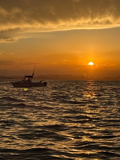 The image depicts a marine scene at either sunrise or sunset, with the sun low on the horizon, casting an intense orange and amber glow across the sky and water. The location is off the coast of Port Kembla, Australia, with the city's industrial complex visible as a silhouette on the distant horizon.

In the foreground and mid-ground, a body of water exhibits moderate chop, reflecting the sun's light in a prominent golden path extending from the horizon towards the viewer. A white motorboat, approximately 20-25 feet in length, floats on the water to the left of the sun's reflection. This vessel features a forward cabin or console area with a dark-colored bimini top. Several fishing rods are mounted vertically on the boat, and an outboard motor is visible at the stern. A partial numerical registration "4180" is discernible on the port side hull. A distinct, diffused yellow-green light or reflection is visible just beneath the water's surface, adjacent to the boat's bow. One human figure is partially visible within the boat's cabin area.

The background reveals the industrial skyline of Port Kembla, characterized by multiple factory structures, tall chimneys, and cranes, all rendered in dark silhouette against the bright sky. A plume of white smoke or steam is observed rising from at least one of these industrial structures. Low-lying clouds are present above the industrial complex and partially obscure the sun. The upper portion of the sky is dominated by elongated, dark-edged clouds against a lighter orange-yellow hue.