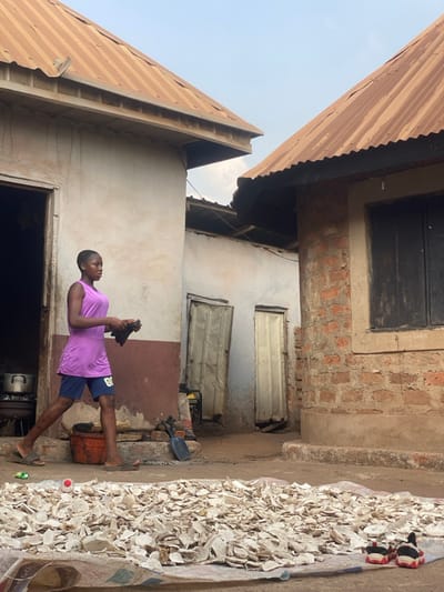 The scene depicts an outdoor courtyard in Aliade, Nigeria, surrounded by multiple residential structures under a clear sky. A young, dark-skinned individual with short hair, wearing a purple sleeveless top, dark blue shorts, and sandals, walks from left to right across the mid-ground. She holds a dark, possibly fabric, object in her right hand.
In the foreground, a large pile of irregularly shaped, light-colored dried material, likely cassava chips, is spread on a light-colored tarp or sack on the unpaved ground. A small red and white spherical object rests atop the pile. To the lower right of the pile, a pair of small black sneakers with red soles are placed.
To the left, a single-story building with a light-colored facade and a dark reddish-brown base has an open doorway revealing stacked metallic pots within. Its roof is constructed from rusted corrugated metal. Directly opposite, to the right, stands another single-story building with exposed reddish-brown brickwork and a dark, possibly shuttered, window. This building also features a rusted corrugated metal roof. In the background, between these two primary structures, two more buildings with light-colored walls and worn, corrugated metal doors are visible. A yellow and black object, possibly a small generator, is positioned near the background doors. An orange bucket and a shovel are discernible near the base of the left building.