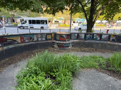 The image captures a public space in Chicago, United States. In the foreground is a round, multi-tiered mosaic-covered structure, encircled by a black concrete wall. The mosaic features various designs, some resembling urban landscapes and industrial motifs. The space within the structure contains a variety of green plants and grasses.
Behind this structure, a sidewalk runs parallel to a street. Several people are walking on the sidewalk and visible at the edge of it and in the background. A white Pace bus is driving on the street, and a white car is parked. A red fire hydrant is located on the sidewalk near the street. The background includes trees and buildings, suggesting an urban environment with commercial establishments. The overall lighting suggests it is daytime.