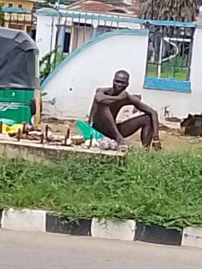A bare-chested man is sitting on the ground with his legs crossed and arms resting on his knees. He is smiling at the camera. Behind him, there is a green tarp and some other objects, possibly for sale. To the left, part of a green and grey vehicle, possibly a tuk-tuk, is visible. In the background, there is a white building with blue trim and barred windows, and some trees. The scene appears to be outdoors, in what looks like a marketplace or roadside setting. The lighting suggests it is daytime, possibly overcast. The man's expression is one of apparent contentment or amusement. There is no visible text in the image.