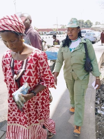 An outdoor scene in Communauté rurale de Payar, Senegal, depicts a street environment under bright daylight. In the foreground, two women walk from left to right on a paved sidewalk. On the left, an older dark-skinned woman with a serious expression looks downwards. She wears a red and white patterned traditional dress featuring geometric triangles on the upper portion and vertical stripes on the lower. A matching striped headwrap covers her head. She displays gold hoop earrings and a dark bracelet on her right wrist, which holds a substantial stack of banknotes. A black and white patterned strap from a shoulder bag is visible over her left shoulder. She wears light-colored sandals.
To her right, a younger dark-skinned woman with braided hair looks forward and slightly left. She wears a light green, long-sleeved uniform consisting of a collared shirt with breast pockets and cargo pants, paired with orange work boots featuring black laces. A white t-shirt with a green V-neck and a partial "VC" or "YC" logo is visible underneath her uniform shirt. She wears a matching light green cap with a white and green logo. A small black messenger bag hangs over her left shoulder, and she holds a small white paper or card in her right hand.
In the mid-background, partially obscured by the foreground woman, a bald dark-skinned man in a maroon shirt is visible, looking right. Behind him, a market stall with assorted goods, possibly produce, covered by a light blue tarp, is attended by another individual. The background reveals a street with multiple cars, some parked and others potentially in motion, separated from the sidewalk by a curb and a median strip marked with dark bollards. A low reddish-brown wall or building is visible in the far background under a clear sky. Debris is visible along the edge of the sidewalk on the right.