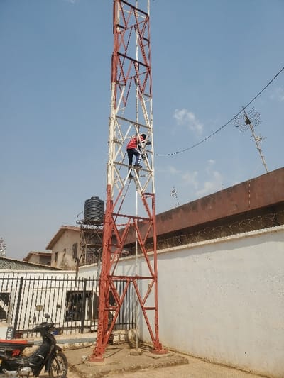 This image, captured in Sabon Gari North, Nigeria, depicts an outdoor urban scene featuring a communications tower and a person working on it under a clear sky.

**Central Elements:**
A lattice communication tower, painted in alternating sections of reddish-brown and off-white, dominates the center-right. Approximately one-third of the way up the tower, an individual is observed. This person wears a red long-sleeved top over a dark undershirt or garment and dark trousers, appearing to be actively engaged with equipment or cabling on the tower structure. A black cable extends horizontally from the tower towards an adjacent structure.

**Surroundings and Objects:**
To the right of the tower, a white-stuccoed perimeter wall with a dark brown coping is visible, topped with multiple strands of coiled barbed wire. Beyond this wall, an antenna (likely a television antenna) is mounted on a pole on a rooftop. To the left of the tower, a black motorcycle is parked on a paved area. Behind the motorcycle, a metal gate with vertical bars partially obscures a building with a white facade and a dark, possibly industrial, structure. Further back, another light-colored building with a dark roof is visible. A black plastic water tank, elevated on a metal stand, is situated behind the tower and to its left. The ground around the tower base and motorcycle consists of concrete and packed dirt. The sky above is predominantly clear blue with minimal scattered clouds.