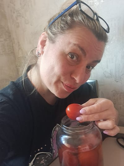 A female subject is depicted in a medium-close, front-facing perspective within an indeterminate domestic interior in the city of Меткомелино, Russia. The subject has light-colored hair, pushed back, with dark-framed eyeglasses with blue accents resting on her forehead. She wears a dark-colored t-shirt, a thin chain necklace, and a silver-toned earring with a red stone in her left ear. Her left hand, with light pink nail polish, holds a single red, glossy tomato directly above the opening of a clear glass jar. The jar is partially filled with similar red tomatoes submerged in a reddish liquid. The subject's right hand is partially visible, showing a thin gold-colored ring on the ring finger. She is looking directly into the camera with a slight smile and her lips slightly parted. The background consists of a light-colored wall covered with patterned wallpaper featuring faded, abstract designs. A pair of black-handled scissors is partially visible on a light-colored surface to the right of the jar.