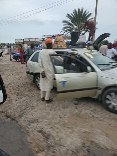The image depicts a street scene in Dangar, Nigeria, on a cloudy day. In the foreground, a beige sedan is parked with its passenger door open. A man in a light-colored traditional outfit stands next to the open door, facing the car. Behind the sedan, a blue vehicle is parked, and several people are visible around it. To the left, a gas station with the signage "ALH. HAMIDU MADU" is present. In the background, a tall palm tree stands prominently. The ground is unpaved and appears to be a mixture of dirt and gravel. The overall impression is one of activity and movement, possibly related to travel or commerce.