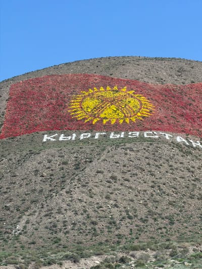 This is an aerial, slightly distorted view of a mountain slope in Kyrgyzstan, featuring the flag of Kyrgyzstan painted onto the rock face. The flag consists of a red background with a yellow sun emblem in the center. To the right of the flag, written in white letters, is the word "КЫРГЫЗСТАН" (Kyrgyzstan) in Cyrillic script. The mountain slope is arid and rocky, with sparse green vegetation. A thin black cable, possibly part of a ski lift or power line, runs diagonally across the mountain face in the upper right portion of the image. The sky is a clear, bright blue. The lighting suggests it is daytime, likely late morning or early afternoon, with no clouds visible. The scene appears peaceful, with the prominent display of national symbols on the natural landscape.
