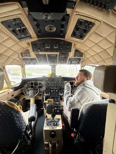A man with a beard, wearing a gray hoodie, is seated inside the cockpit of an aircraft. The cockpit is filled with various panels of buttons, switches, and gauges. The interior is upholstered with quilted beige padding. Through the aircraft's windshield, a rainy outdoor scene is visible. The exact location within Kraaifontein, South Africa, cannot be determined from the image.