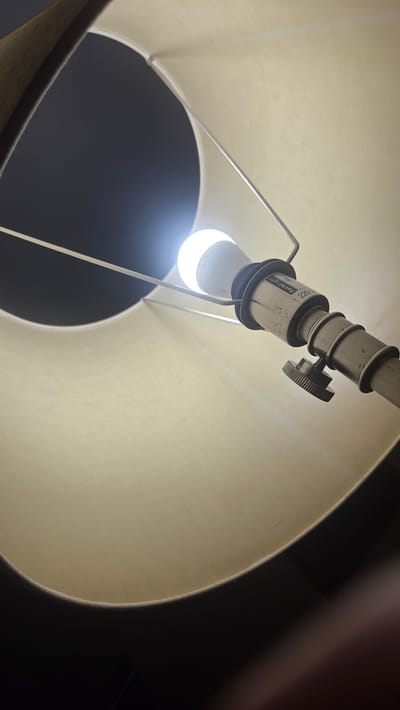 The image is a close-up, low-angle shot from inside a lampshade, looking upwards towards the light bulb and its fixture. The lampshade itself appears to be made of a light-colored fabric, possibly cream or beige, with a textured surface. The interior of the lampshade is illuminated by a bright, white LED light bulb. The light bulb is screwed into a metal fixture, which is supported by thin metal arms extending from the top rim of the shade. Part of the lamp's pole, with a textured knob, is visible on the right side of the image. The background outside the lampshade is dark, suggesting it is nighttime or the room is dimly lit. No specific location in Bray, Ireland can be identified from this image.