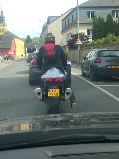 A person is riding a Suzuki motorcycle, bearing license plate XW 7749 from Luxembourg. The motorcycle is traveling on a street in Fentange, Luxembourg. The vehicle is traveling past residential buildings. Parked vehicles are to the right. The photo was taken from another vehicle.