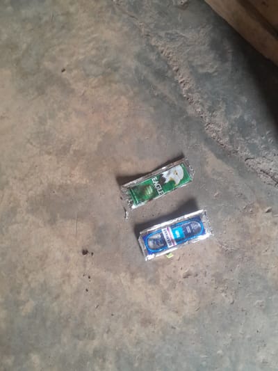 The scene is located in Ifo, Nigeria. Two small, rectangular packaged items rest on a dusty, mottled concrete surface. The upper item is a green packet, partially crumpled, bearing the brand name "EAGLE" and an eagle head logo. Below it, the second item is a blue packet, also partially crumpled, with the visible brand names "SEAMAN'S" and "DAILY" alongside a fish graphic. The ground is a light greyish-brown, uneven, and shows evidence of wear, dirt, and small debris. A dark, out-of-focus object is visible along the top right edge. No individuals or actions are depicted.