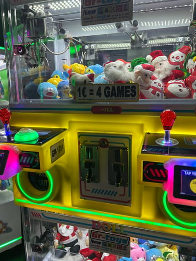 This media file captures a brightly lit arcade claw machine, likely situated in an amusement center or similar commercial establishment in Agios Dimitrios, Greece. The dominant subject is the machine itself, which is largely yellow with vibrant neon green, pink, and blue lights emanating from its control panel and edges.

Inside the clear glass casing, numerous plush toys are visible, including several Santa Claus figures, a white fluffy toy, blue cartoon characters resembling Doraemon, and other assorted small stuffed animals like a white cat-like figure.

The machine's control panel features a prominent red, textured joystick with a clear knob, surrounded by a glowing green ring. Below the joystick, two coin slots or bill acceptors are recessed into the yellow casing. A digital screen on the control panel displays partial text, including "TAP YO", suggesting interaction prompts. A prize chute labeled "PRIZE OUT" is visible towards the bottom right of the panel.

Several stickers with text are present. One notable sticker inside the machine states "1€ = 4 GAMES", indicating the cost and number of plays. Another sticker reads "DOLL". Manufacturer or operator information is displayed on white rectangular stickers, including "Gr-toys New", the website "www.Gr-toys.gr", and a phone number "210 44 17 208". Greek text "(ΠΩΛΗΣΕΙΣ - ΣΥΝΕΚΜΕΤΑΛΛΕΥΣΕΙΣ - SERVICE)" which translates to "SALES - JOINT VENTURES - SERVICE" is also visible. The machine itself is labeled "GR-1784". The claw mechanism is suspended over the toys, ready for play. The image is taken from an elevated angle, looking down into the machine.