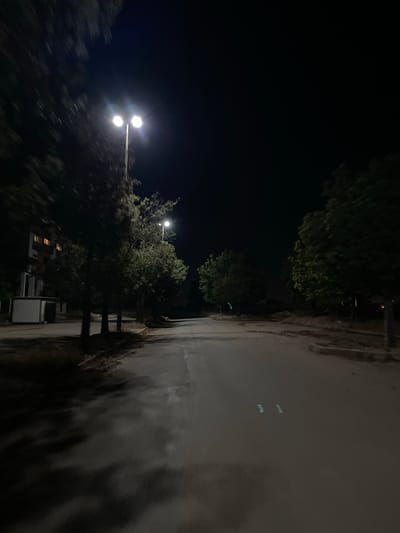 At night in Dar Oum Soltane, Morocco, a street is illuminated by streetlights. Trees line the side of the road. A building is visible in the distance. The street surface appears wet and reflective.