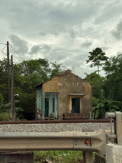 An abandoned, weathered, yellow-hued single-story building stands beside a railway track in the Hải Lăng Commune of Vietnam. The building shows significant disrepair, with a partially collapsed roof and peeling paint. The entrance features green doors and ornate window grates, hinting at former habitation. In front of the building, a stack of weathered wooden planks rests near the railway. Lush green foliage, including banana trees, surrounds the structure. A utility pole with wires stands to the left, and a grey metal guardrail is prominent in the foreground. The sky is overcast with heavy clouds, suggesting a gloomy atmosphere. There are no people visible in the image.