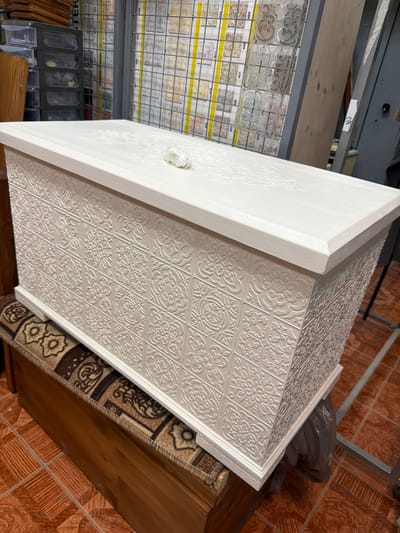 This media file depicts a close-up, slightly angled view of a white, embossed cabinet or chest. The cabinet's surface features a repeating pattern of decorative, floral-like motifs within square panels. The embossing creates a textured, three-dimensional effect. The cabinet appears to be made of a light-colored material, possibly wood or MDF, and has a clean, unfinished look on its left side, revealing a slightly textured grain.
In the background, shelves are visible, laden with what appear to be samples of tiles or flooring materials. These samples are arranged in grids and display various colors and patterns, suggesting a retail or storage environment, possibly a hardware store or showroom. Some of the sample cards have printed numbers and text, such as "409," "408," "407," "050," and "049."
To the right of the cabinet, a brown, wooden surface is partially visible, with a patterned rug or fabric laid on top. The rug has intricate designs in brown, beige, and black. The floor in the background is tiled with reddish-brown, square tiles arranged in a diamond pattern. A crumpled dark plastic bag is also visible in the upper right background.
The lighting suggests an indoor setting, likely during daylight hours, though no windows or natural light sources are clearly visible. The image is oriented in a way that suggests the cabinet is positioned on a floor or low surface. There are no people or animals visible in the frame. The overall impression is one of a product display or storage area within a commercial space. The location context is identified as Maloyaroslavets, Russia.