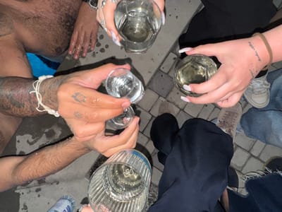 The image provides an overhead view of at least four individuals engaged in a social gathering in Yeghvard, Armenia. The scene appears to be set outdoors, possibly at night or in low light, near a body of water.
**People:**
* **Upper Left:** A male with a hairy torso and arm is visible, appearing wet and positioned near a blue water body, likely a pool. His right hand holds a clear drinking glass.
* **Lower Left:** A male with tattooed forearms and a white shell bracelet holds two clear shot glasses in a raised position, as if toasting. His hands are wet.
* **Upper Right:** A female's left hand, with long, light-colored manicured nails and multiple rings, holds a large clear drinking glass.
* **Middle Right:** A female's right hand, featuring a forearm tattoo (text and dots) and a brown wristband, holds a large clear glass containing a light yellow liquid. Her lower legs are visible, dressed in light-colored sneakers and blue jeans.
* **Lower Right:** Additional individuals are present, evidenced by visible lower legs in dark pants and blue jeans with distressed hems, alongside various casual footwear on a paved surface.
**Objects:**
* Five clear drinking glasses are central to the composition: three larger tumblers (two containing light yellow liquid, one with clear liquid) and two smaller shot glasses (both containing clear liquid).
* A partially visible blue plastic water bottle is on the ground in the lower left.
* Water droplets are visible on hands and the surrounding ground.
**Actions/Interactions:** The individuals' hands are raised, holding their respective glasses, indicating a shared toast or communal drinking activity.
**Environment:** The ground consists of grey or light-colored interlocking pavers. A visible blue body of water, likely a pool, is situated on the left side of the frame.