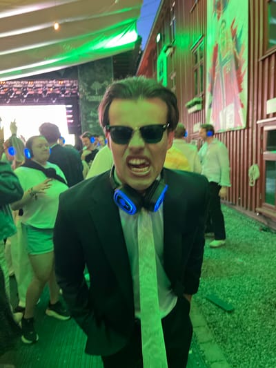 The image captures a close-up of a young man wearing sunglasses, a suit jacket, and a tie, with blue-lit headphones around his neck. He is in a crowded outdoor setting, possibly a festival or event, with a blurred background showing other people also wearing headphones. The environment is illuminated with green lighting, and a wooden building with a large banner featuring a person in athletic wear is visible on the right. The ground appears to be covered in gravel. The person in the foreground is making a loud expression, with their mouth open. The presence of a banner with a Norwegian flag suggests the location is in Norway, and potentially Tønsberg, Norway, if that is indicated by other visual cues or external information not present in the image itself.