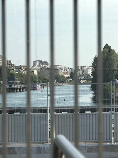 The image shows a reflection of a city skyline, a river, and a tree, seen through horizontal bars. The buildings are modern, with white and grey facades. A red and white boat is visible on the river. The reflection is distorted by the bars, creating a segmented view of the scene. The sky is overcast, suggesting a cloudy day. The location appears to be an urban setting, likely along a river or canal, given the presence of the boat and the cityscape. The time of day is difficult to determine precisely, but the lighting suggests it is daytime. No people are clearly visible, but the scene implies human activity and urban life. The reflection also shows a large, green tree. The overall mood is calm and somewhat abstract due to the bars obscuring parts of the image.