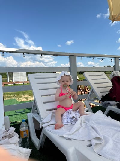 An infant wearing a pink swimsuit and a white sun hat sits on a white lounge chair. A pacifier is in the infant's mouth. The infant is holding a small object in their hands. A white towel is draped over the lounge chair. In the background, there is a wooden fence, green grass, and a building with white siding. String lights are visible overhead. A plastic bottle of water is on the ground next to the lounge chair. The scene appears to be an outdoor recreational area, potentially near Колокша, Russia, given the reference. The sky is blue with scattered white clouds.