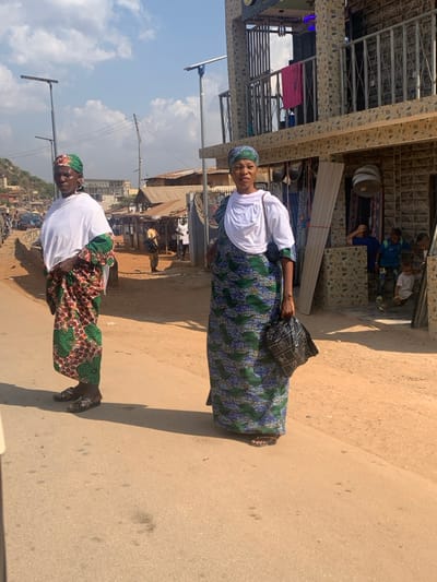 The image depicts an outdoor street scene in Egbedi, Nigeria, under bright daylight with a clear sky and scattered clouds. In the immediate foreground, two adult women of dark complexion are standing on a light-colored asphalt road. The woman on the left wears a white short-sleeved top over a long, patterned wrapper featuring green, red, and brown motifs, with a matching headwrap and dark sandals. The woman on the right, positioned slightly closer to the camera, wears a white short-sleeved top with a white cloth draped over her left shoulder, a long patterned wrapper in blue and green, a matching headwrap, and dark sandals. She holds a black plastic bag in her left hand.
To their right, the road transitions to a light brown dirt shoulder leading to a two-story building with a textured stone-tile facade in beige and brown tones. The ground floor features an open entrance with various items hanging, possibly merchandise, and a stack of light-colored panels leaning against the wall. Several children are visible near this entrance, some crouching or sitting. The upper floor of the building has a balcony with metal railings, from which a red cloth is hanging, and a sign partially reading "NEW AB..." is visible above.
In the mid-ground, a row of single-story structures with corrugated metal roofs lines the left side of a dirt path parallel to the road. Further down this path, several blurred figures are present. Tall, grey solar-powered streetlights are visible, with one positioned behind each woman. Overhead power lines and poles crisscross the sky. In the distant background on the left, a gentle hill or elevated terrain is discernable. The sun casts distinct shadows to the left and slightly forward of the women.