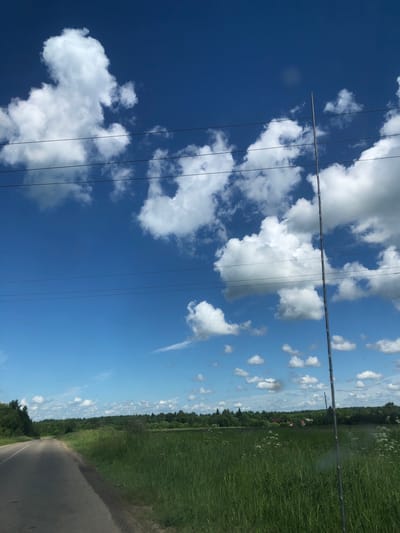 The image depicts a rural landscape in Izdeshkovo, Russia, under a bright blue sky dotted with fluffy white clouds. A paved road curves to the left, bordered by a vibrant green grassy field on the right. In the distance, a dense line of trees forms the horizon, with hints of red-roofed buildings peeking through the foliage. A tall, slender pole, possibly an antenna or utility pole, stands vertically on the right side of the frame, with power lines stretching horizontally across the sky. The scene conveys a sense of openness and natural beauty, characteristic of the Russian countryside.