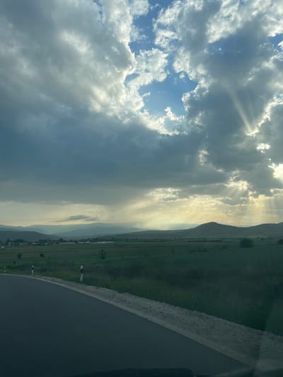 The image shows a view of the Isperihovo landscape in Bulgaria. In the left portion of the image is a cloudy sky with beams of sunlight shining through the clouds. The right portion of the image captures a lush, green field with a mountain range in the background. In the very bottom right of the image is a road.
