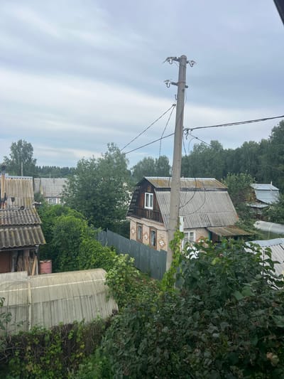 The image depicts a view of a rural area, likely a dacha settlement, in Poletaevo, Russia. The scene is characterized by a collection of residential structures with distinctive corrugated roofs, overgrown vegetation, and a power line pole. A two-story house with a brick first floor and wooden upper section is prominently featured, with a fenced yard in front of it. To the left, another building with a similar roof structure is visible. In the foreground and surrounding the houses, dense green foliage and trees create a lush, natural environment. A greenhouse structure is also present. The sky is overcast, suggesting a cloudy or possibly rainy day. There are no people visible in the image.