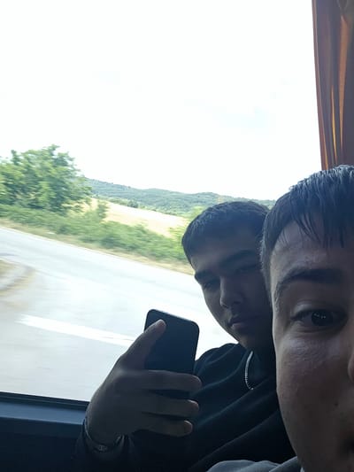 Two young males are captured in a close-up selfie taken from inside a vehicle, possibly a bus, with a blurred landscape visible through the window. The person in the foreground is positioned on the right side of the frame, with their face partially obscured by a close-up shot of their right eye and forehead. They are wearing a dark-colored shirt. To their left, another young male is more fully visible, holding a black smartphone in their left hand, which is positioned in the center of the frame. This individual is wearing a black hooded sweatshirt and a silver chain necklace. The background reveals a road or highway and a rural landscape with green trees and hills under a bright sky. The scene appears to be within or near Krivodol, Bulgaria, suggested by the surrounding natural scenery and the presence of a road, indicative of travel within the region.
