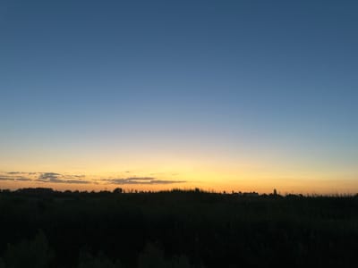 The image captures a serene sunset over a rural landscape in Batash, Uzbekistan. The sky transitions from a deep blue at the zenith to a warm gradient of orange and yellow near the horizon, with faint clouds scattered across the lower portion of the sky. A solitary, distant star is visible in the upper left quadrant of the image. The foreground is dominated by a dark silhouette of dense vegetation, likely reeds or tall grass, creating a stark contrast with the illuminated sky. Beyond the vegetation, a line of trees and some indistinct structures are silhouetted against the sunset. The overall impression is one of tranquility and the quiet beauty of nature at dusk.