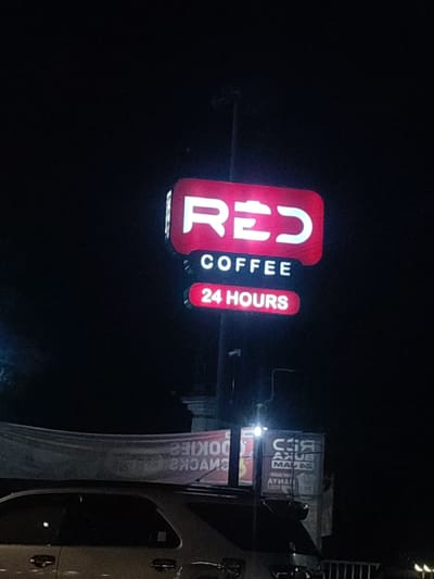 The image shows a sign for a coffee shop called "RED COFFEE" in the city of Kecamatan Pandaan, Indonesia, located at latitude: -7.653 and longitude: 112.688. The sign is red with white lettering and features the words "RED COFFEE" and "24 HOURS" written on separate lines. It is illuminated by a white light and mounted on a pole. The sign is positioned in front of a building, which is partially obscured by darkness. There is a car parked in the foreground with a banner hanging above it, but the text on the banner is illegible. The environment is dark and nighttime. The scene is deserted.