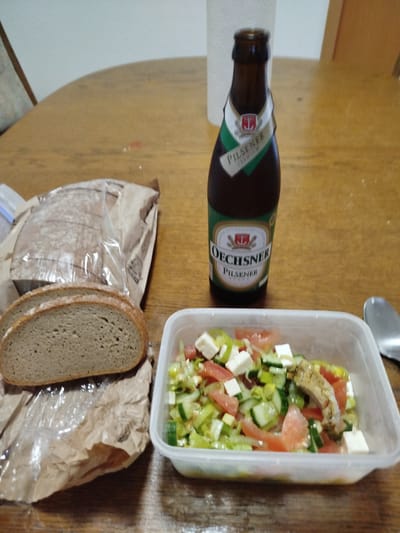 The image depicts a meal laid out on a wooden table. In the foreground, a clear plastic container holds a salad composed of chopped tomatoes, cucumbers, leeks, and cubes of white cheese, possibly feta. A piece of cooked meat is also visible in the salad. To the left of the salad is a loaf of dark bread, partially unwrapped from a paper and plastic bag, with two slices already cut and placed in front of it. Behind the bread and salad, a dark brown glass bottle of Oechsner Pilsener stands upright. The bottle has a label with green and white elements and text indicating "Pilsener Premium". In the background, a white paper towel dispenser is visible, and part of a wooden chair or cabinet. The overall lighting suggests an indoor setting. The scene does not provide any explicit or implicit information about the city of Kitzingen, Germany.