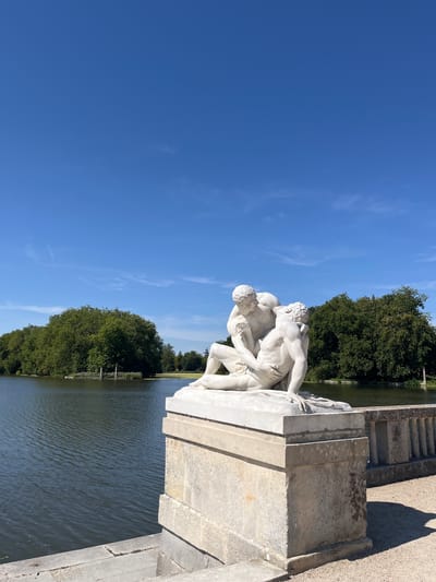 A white marble sculpture depicting two male figures in a dynamic embrace is positioned on a stone pedestal. The sculpture is situated outdoors, adjacent to a body of water, likely a pond or lake, with lush green trees forming a dense backdrop. The sky above is a clear, bright blue with a few wispy clouds. In the foreground, a stone railing runs alongside the water. This scene is located in the gardens of the Château de Rambouillet, in Rambouillet, France. The overall atmosphere is serene and picturesque, indicative of a historical park setting.