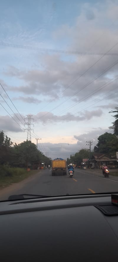 The image shows a view from inside a vehicle, likely a bus or truck, on a paved road. The location is near Paku, Indonesia, inferred from visual cues and the image's origin.

The focus is on the road ahead, where a yellow truck is traveling in the same direction, followed by a motorcycle. A red motorcycle is seen in the distance, moving in the same direction. The road is marked with a yellow line.

To the left of the road, power lines and utility poles are visible against a cloudy sky. Vegetation is present on the roadside. The time of day appears to be dusk or early evening.

The interior of the vehicle is visible on the right side of the image, showing part of the vehicle's wall and a possible window frame.